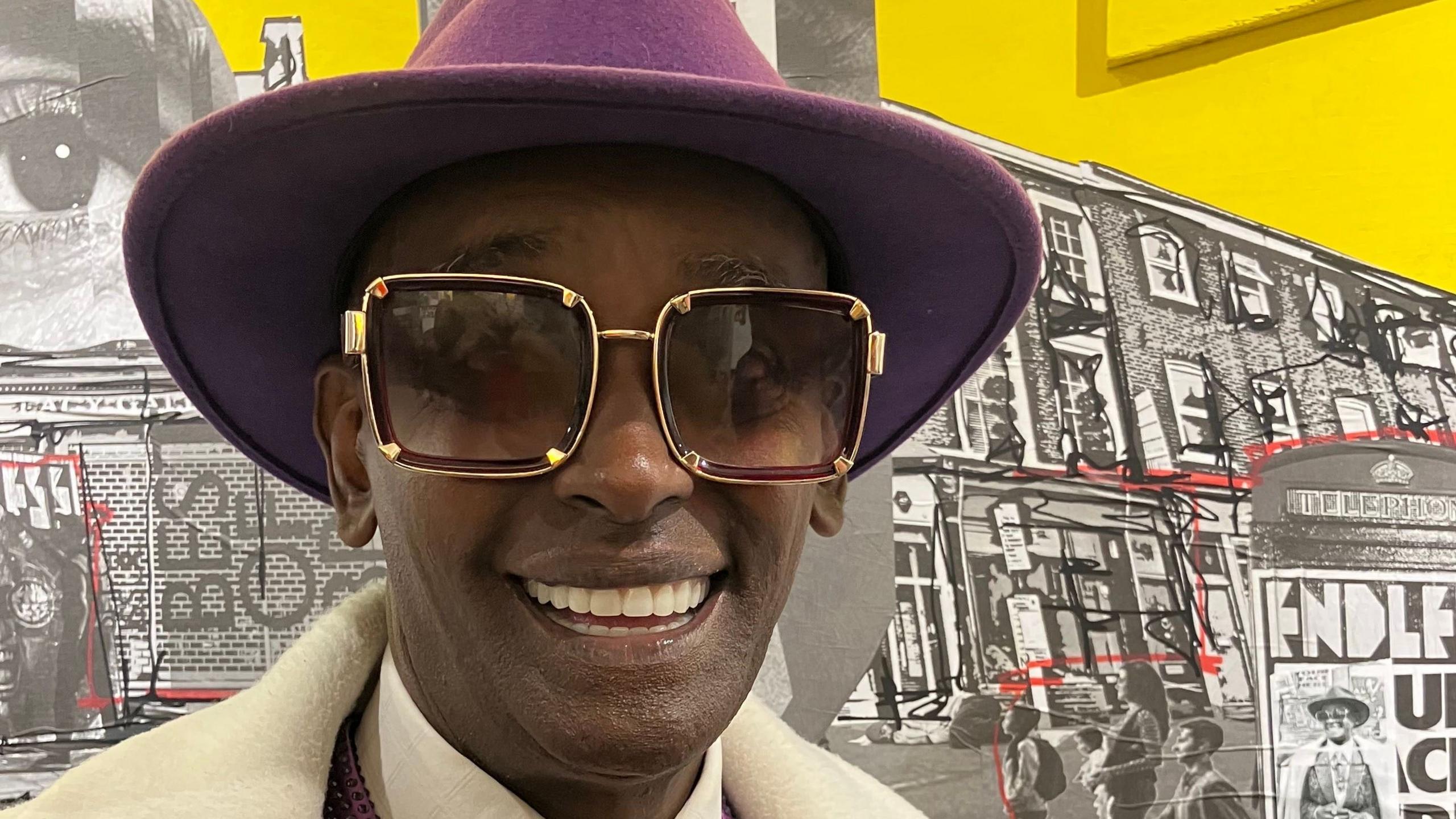 Jama Emli smiles at the camera in a purple suit and sunglasses.
