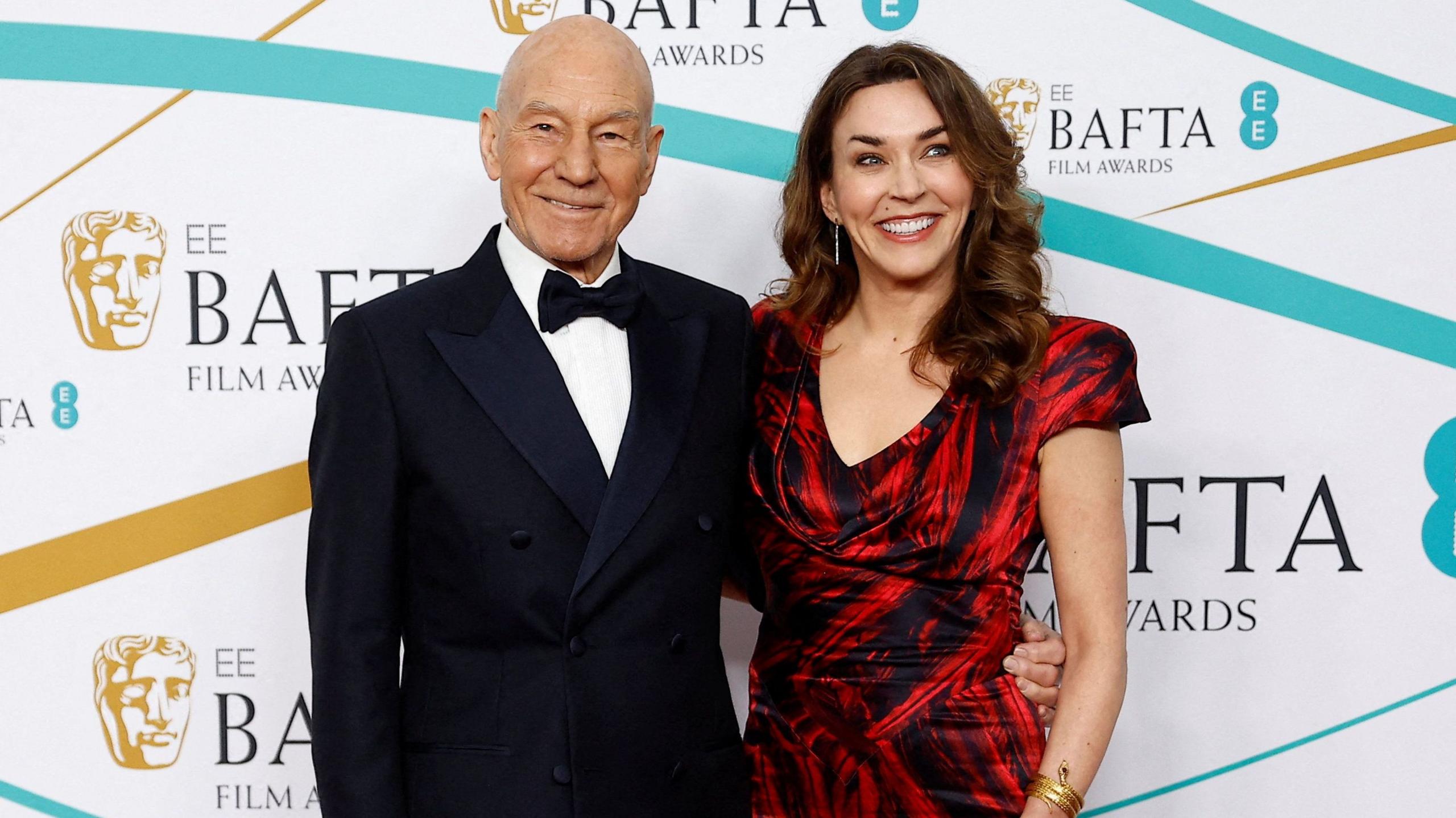 Sir Patrick on the left and Sunny Ozell on the right. He is in black tie and she is in a red and black dress. The background is a the EE Bafta awards backdrop