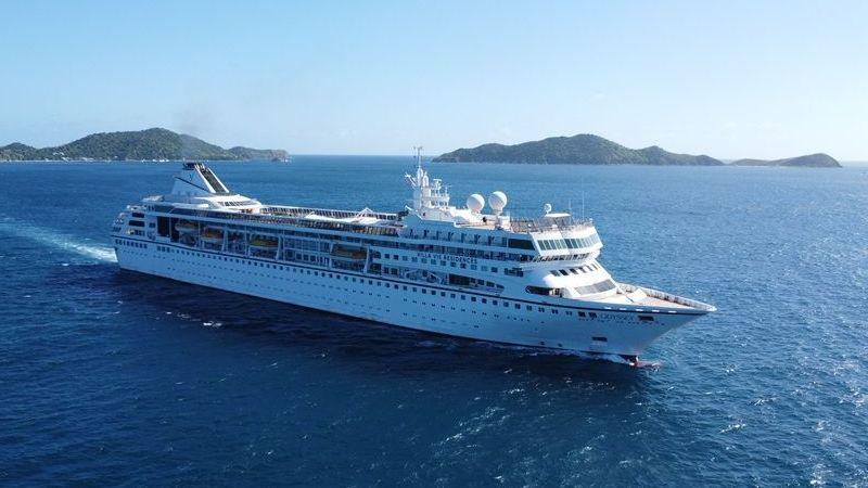 A large white cruise ship sails through deep blue ocean waters near a chain of green, hilly islands under a clear sky.