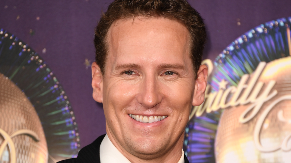 Brendan Cole attends the 'Strictly Come Dancing 2017' red carpet launch at Broadcasting House on August 28, 2017. He wears a black suit and black bow. He has brown hair and stands in front of a Strictly branded purple glitter ball background.