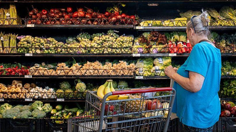 A woman in a blue tshirt stands next to a supermarket trolley as she looks at an aisle stacked with a large variety of vegetables on shelves reaching as high as her head.