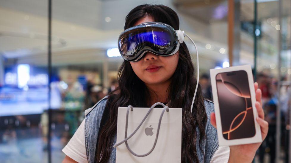 A woman standing in what looks like a bright, modern tech store. They’re wearing a large reflective mixed‑reality headset with a cable trailing from it, the kind that covers the upper half of the face like a visor. In one hand they’re holding an Apple shopping bag, and in the other they’re carrying a boxed iPhone. The background is softly blurred, with glass panels and indistinct figures suggesting a busy retail environment