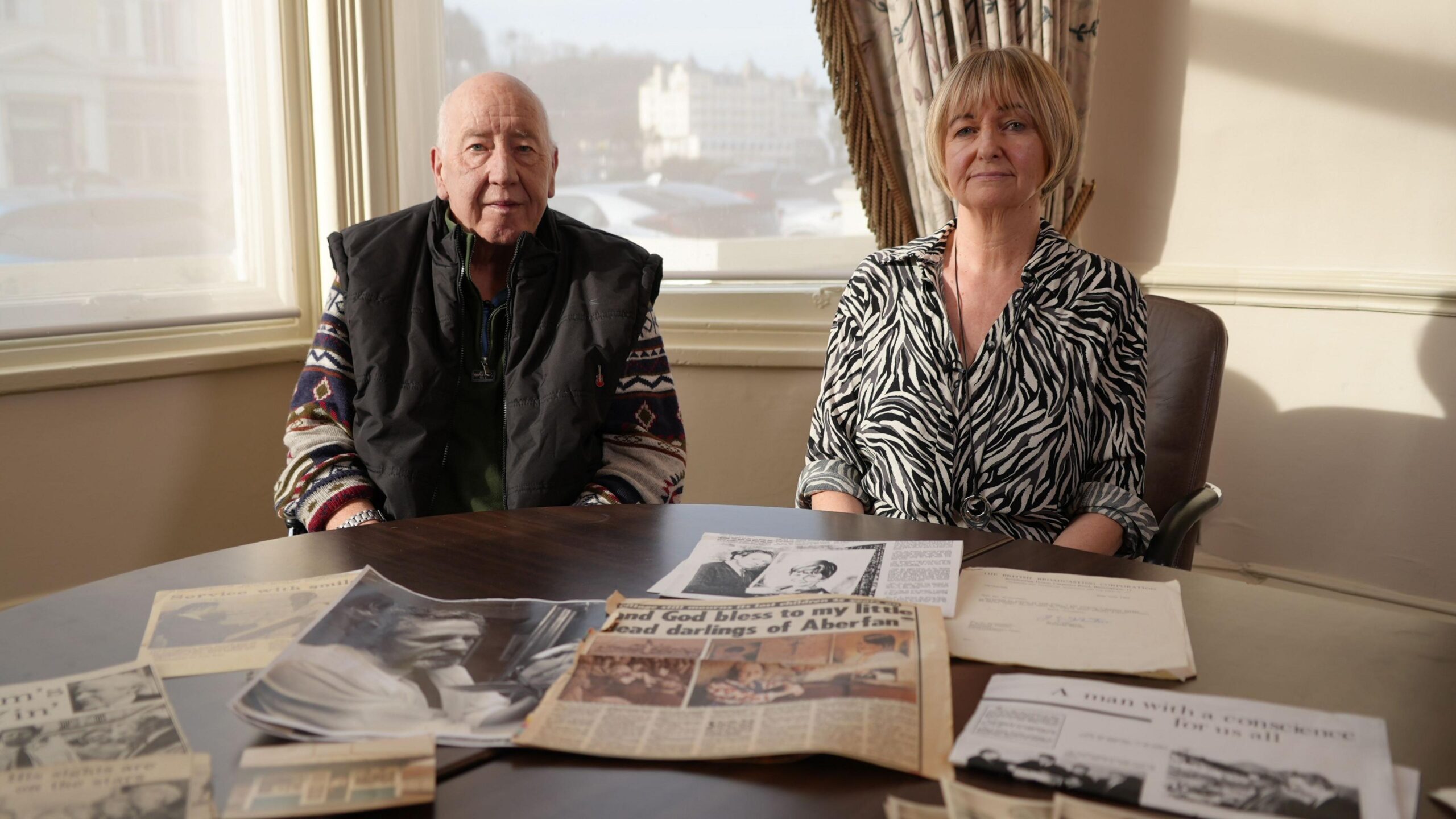 A man and woman sitting at a table that's covered in news paper cuttings and photographs