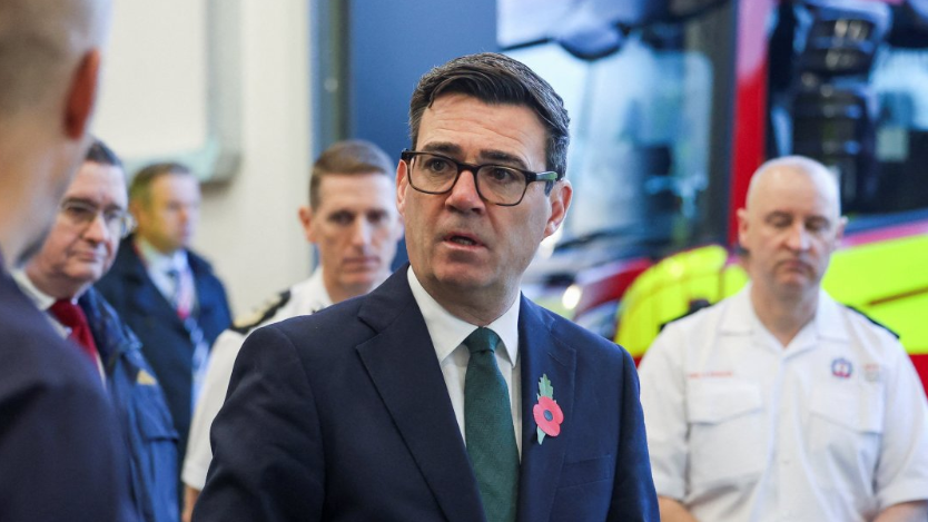 Head and shoulders shot of Burnham meeting firefighters, who are visible in the background along with a fire engine. He is wearing a navy suit, a white shirt and a green tie, with a poppy on his lapel.