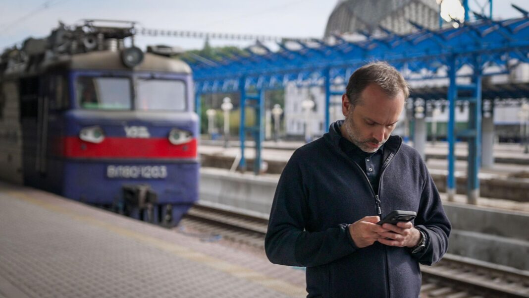 Oleksandr Pertsovskyi, wearing a navy zip up jumper, stands on a platform looking at his phone with a blue and red train in the background