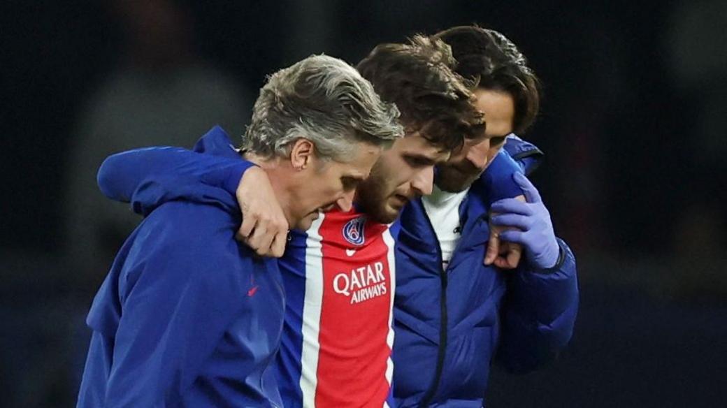 Khvicha Kvaratskhelia (centre, wearing a blue and red PSG shirt) is helped from the field by two men wearing dark blue jackets