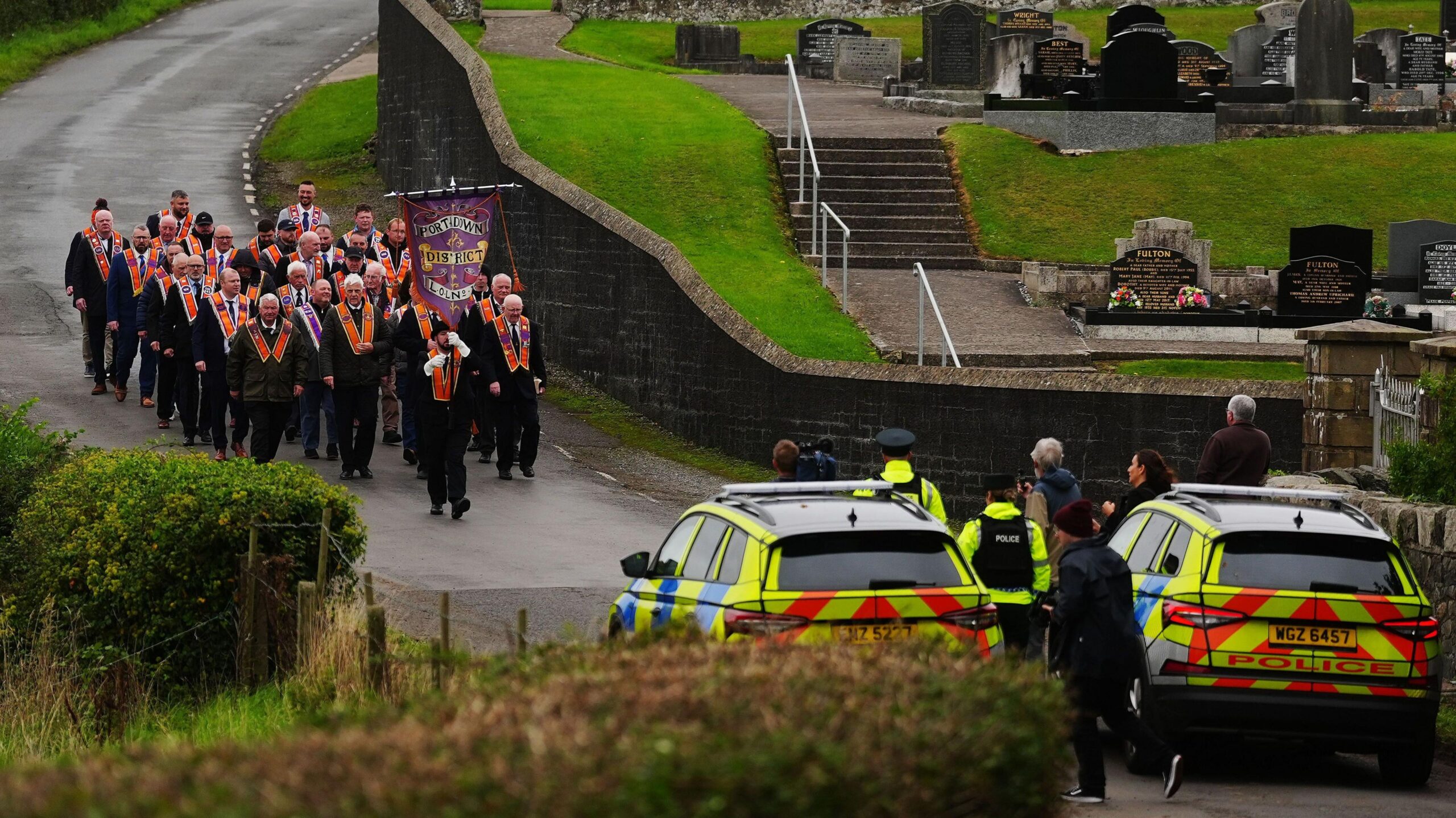 A group of Orangemen walking down a country road. There are two police cars in front of them, blocking their way.
