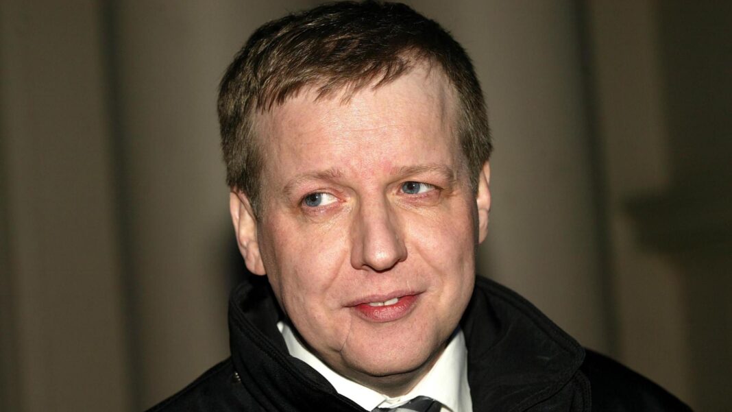 Paul Ferris, a pale man with a scarred face, is wearing a dark coat, white short and dark tie. He is smiling and looks off camera against a curtain backdrop.