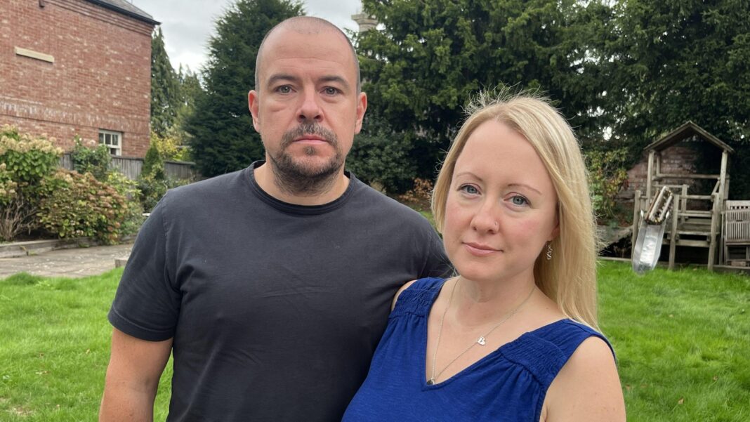 ‘I was blamed for my baby’s death, now NHS needs to change’ Neil Russell has a beard and is wearing a dark t-shirt. Next to him his wife Katie is wearing a blue sleeveless dress and has long blonde hair. They are pictured standing on grass in a garden in front of a brick building.