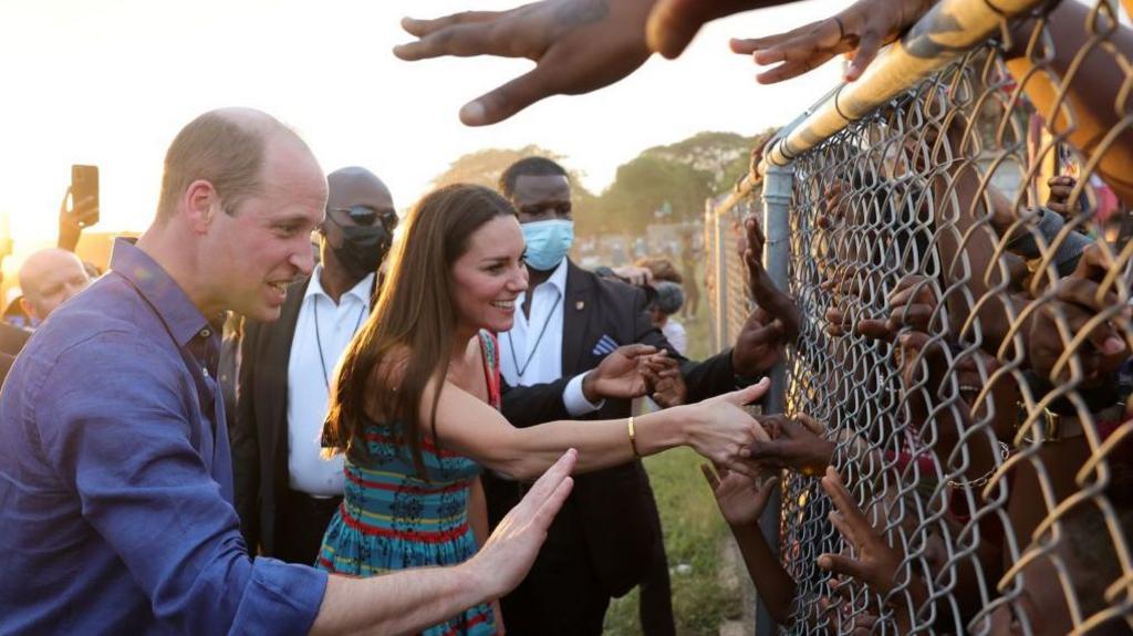 Prince William, Duke of Cambridge and Catherine, Duchess of Cambridge shake hands with children through a wire fence during a visit to Trench Town
