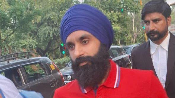 Jagtar Singh Johal looking off camera. He is wearing a red polo shirt with the word 'Boss' on the upper right chest. He is wearing a purple turban around his head and has a long, dark beard. He is walking on a street with cars and green foloage behind him.