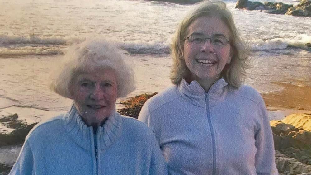 An old photo of Sheila and Bern who are both wearing light-coloured jumpers while standing on a beach with the sea in the background.