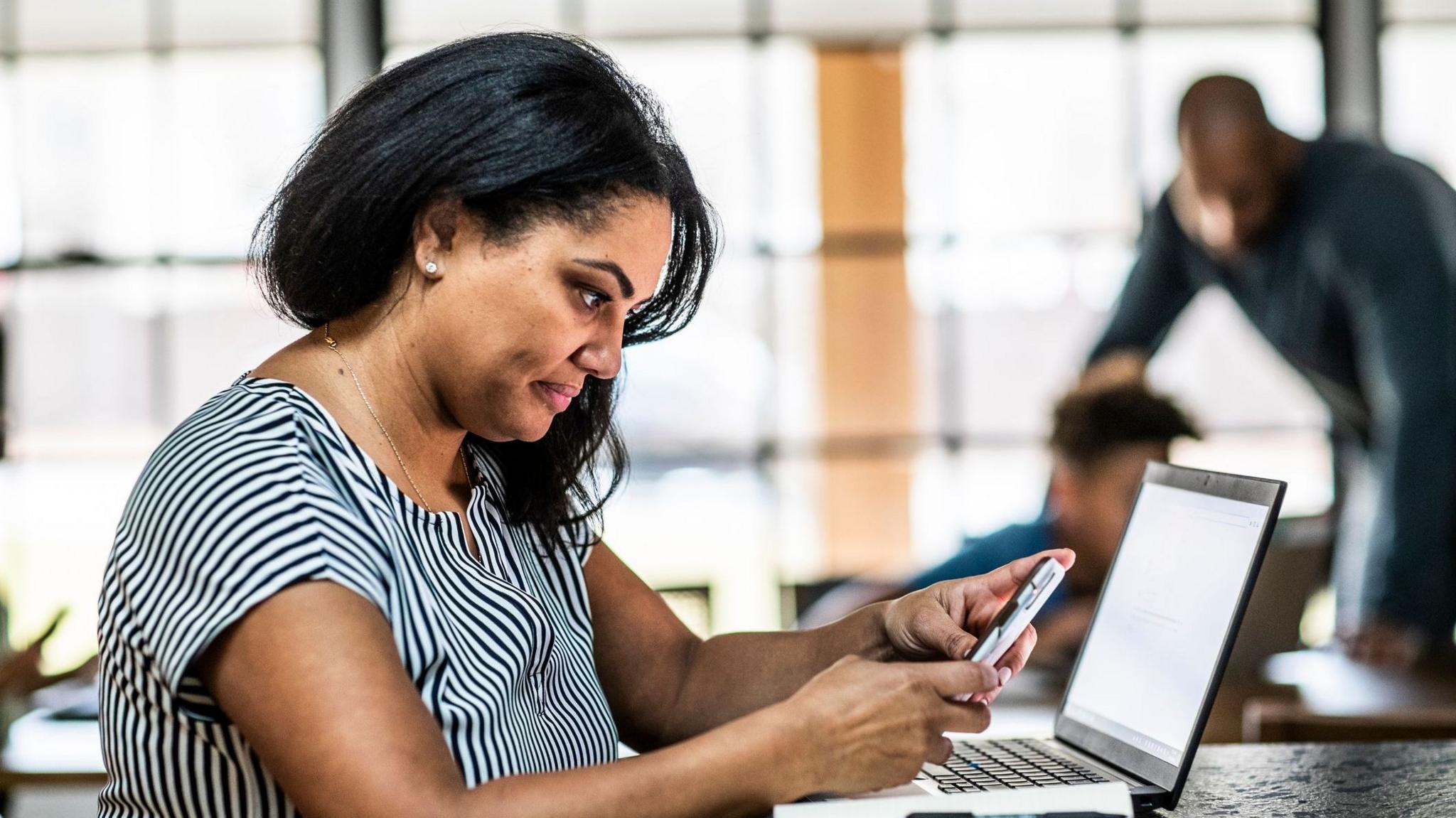 Woman looking at a phone sits at a desk with a laptop in front of her and a man and boy in the background.