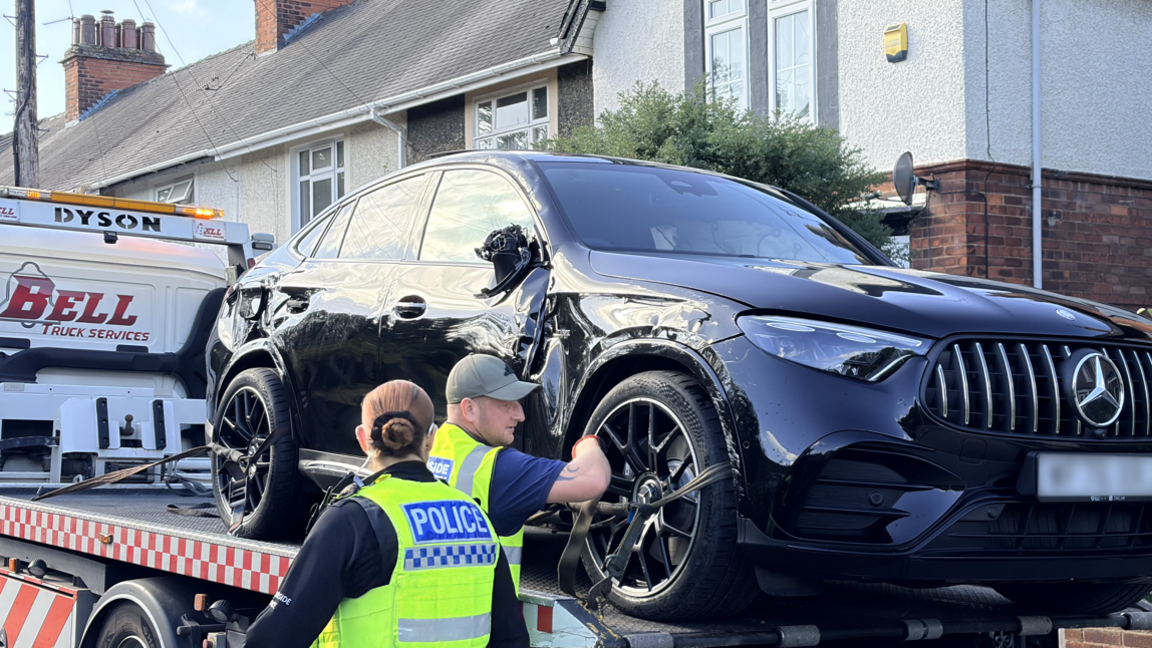 A black Mercedes is being strapped onto a recovery lorry by a worker in a high-vis vest. A female police officer is also standing by.