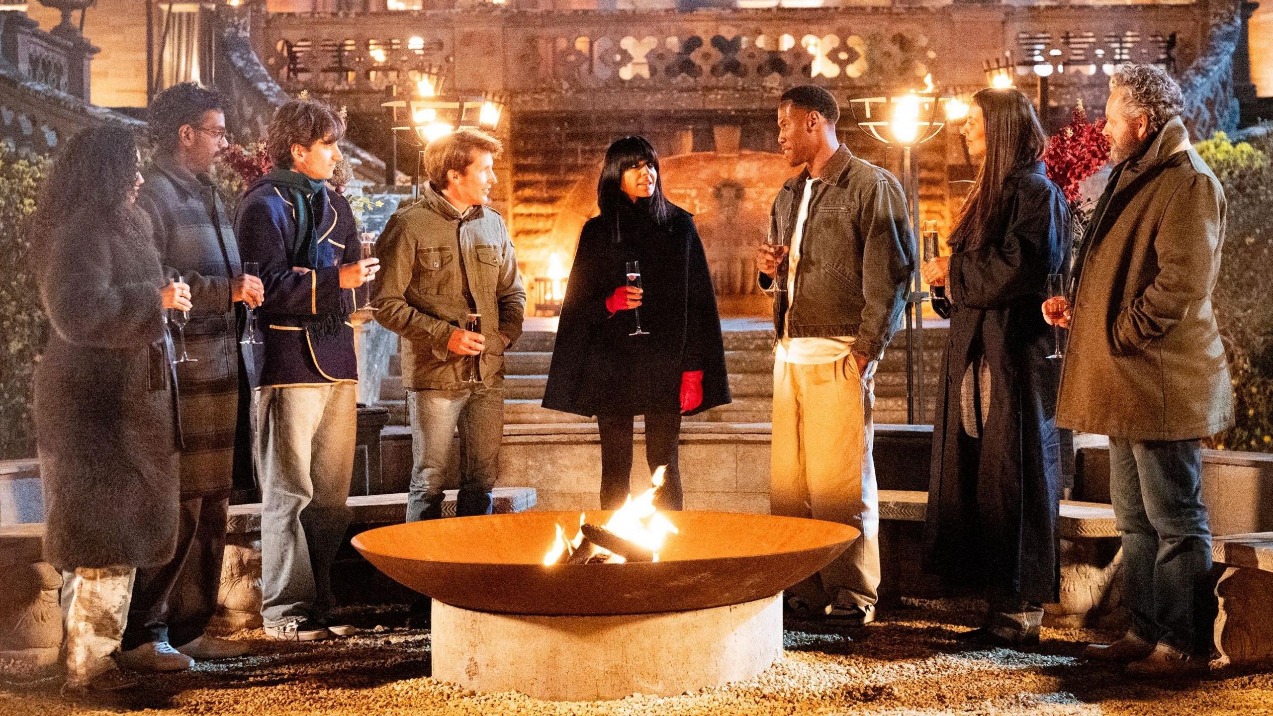 The seven Celebrity Traitors finalists standing around a fire pit outside the castle listening to Claudia Winkleman