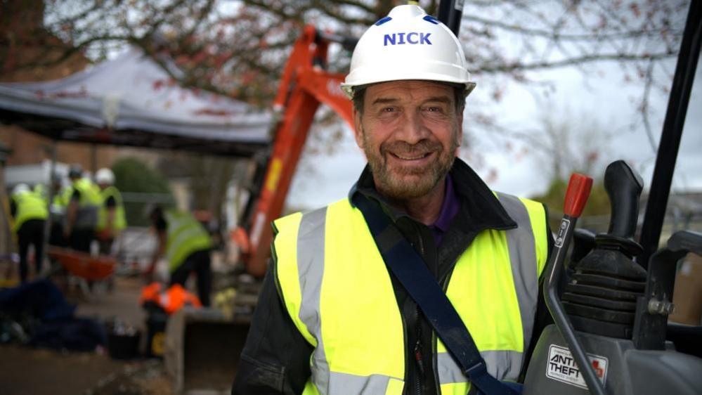 Nick Knowles smiles in front of a building site in a hard hat with his name on it