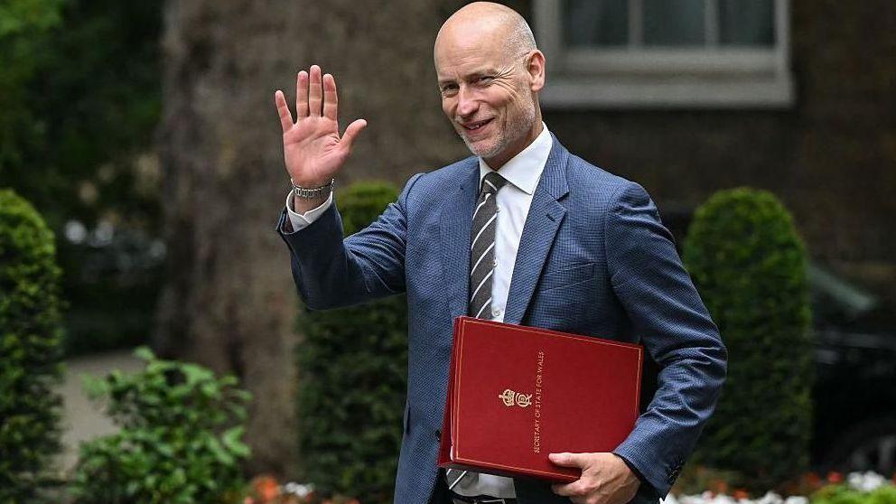 Stephen Kinnock arrives in Downing Street carrying a red Welsh secretary file and raising a hand in greeting. Trees, trimmed shrubs and flowerbeds can be seen in the background.
