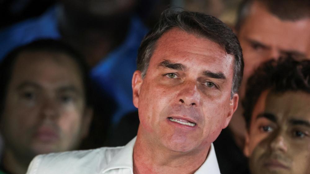Brazilian presidential candidate Flavio Bolsonaro of the Liberal Party speaks after losing the presidential runoff election, in Brasilia on 25 October, 2026. He is wearing a white shirt with an open collar. He is gesturing with one hand. Behind him, his supporters look on.