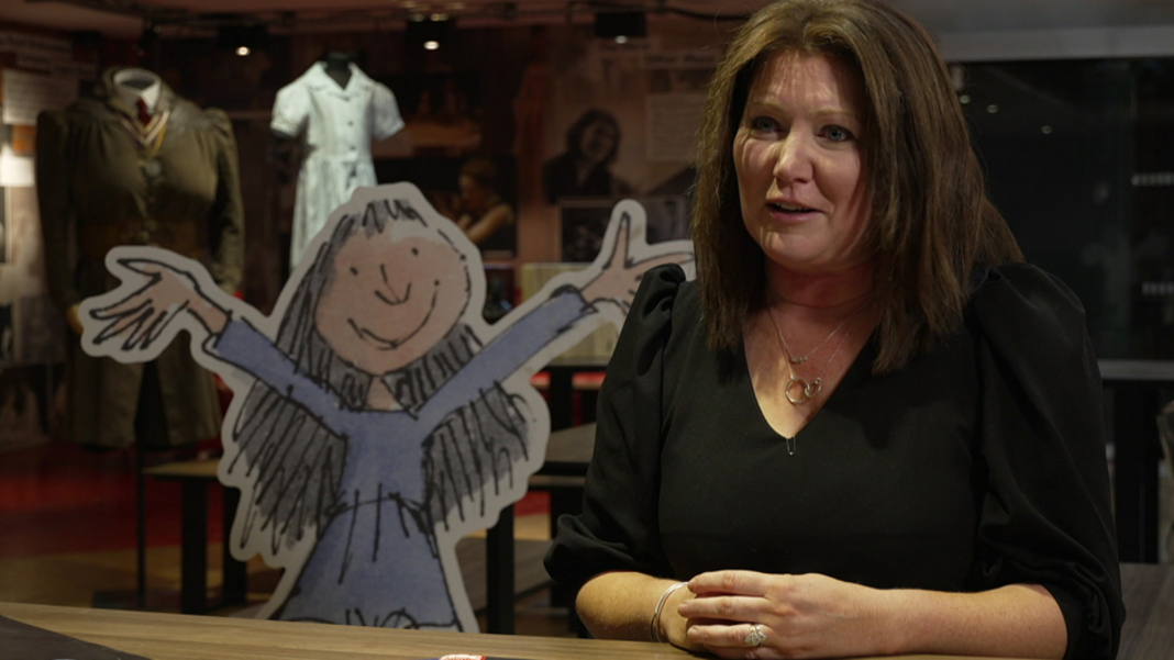 Matilda celebrates 15 years as West End success story Jacqui sits at a desk at the RSC being interviewed. She is looking at the camera and wearing a black top. A cut out of Matilda from Dahl's book stands next to her.
