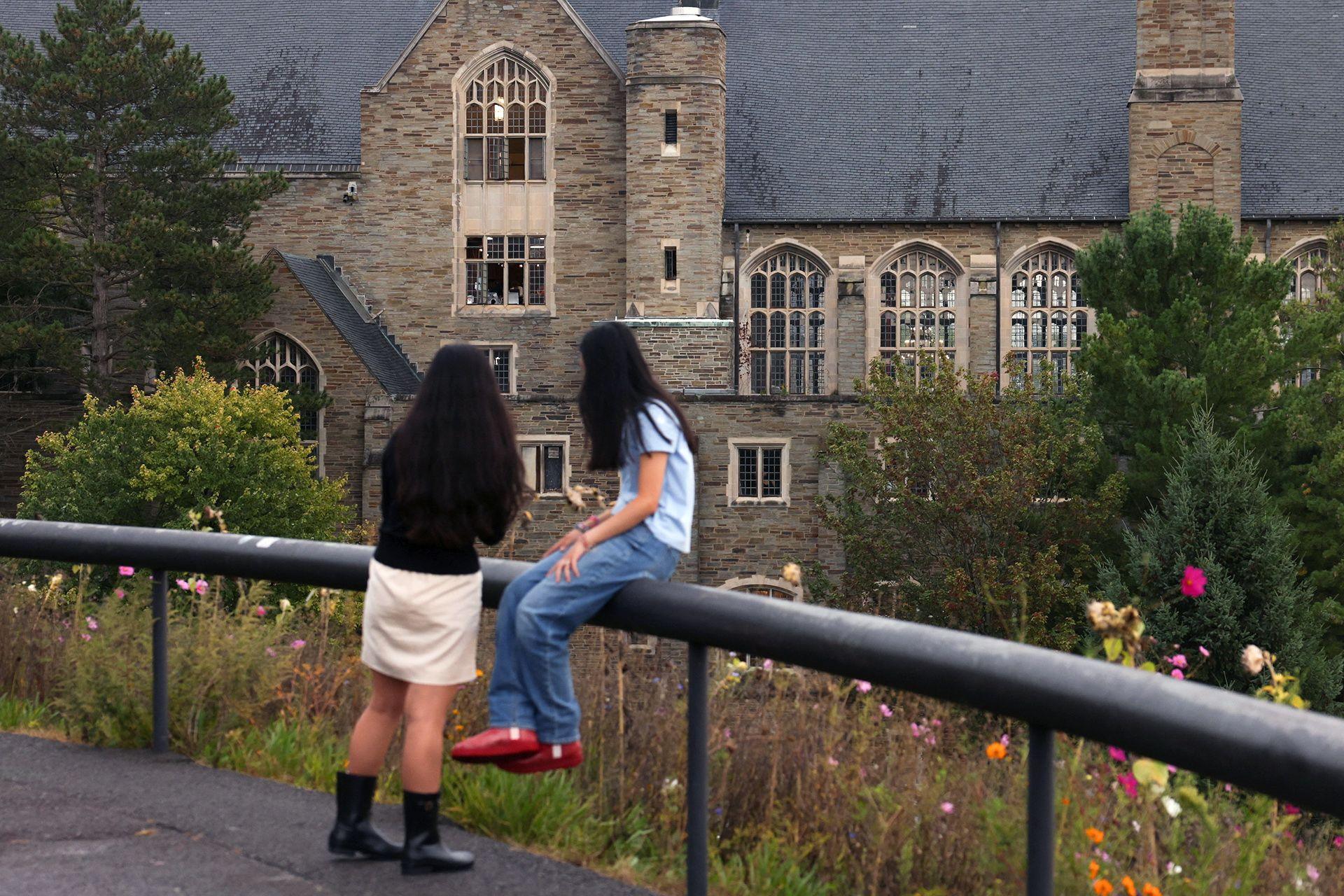 Two students sit on a railing with their backs to camera with the building of Cornell campus in the background amid some tree and flower foliage, taken in Ithaca on Thursday.