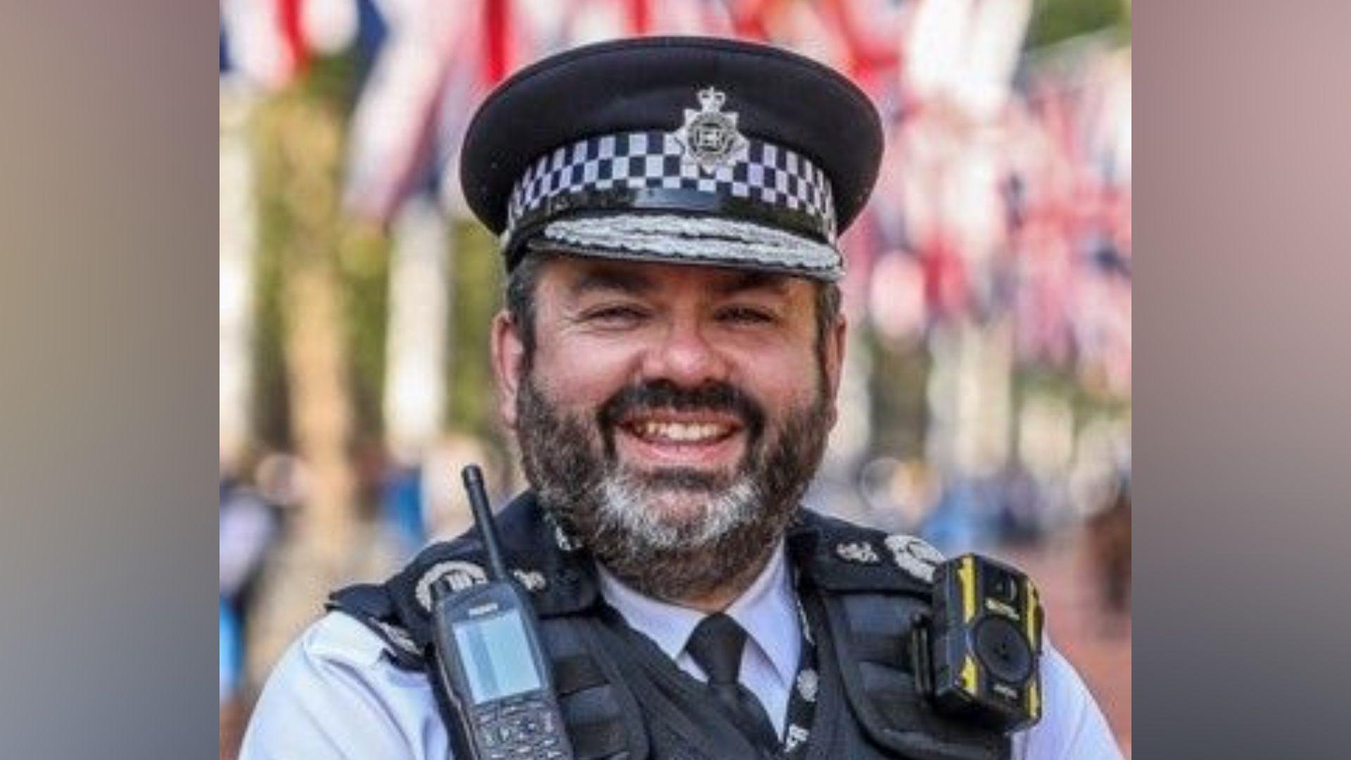 A man in a Metropolitan Police uniform and peaked cap stands outdoors, wearing a radio and body-worn camera on a protective vest. Red, white and blue flags are visible blurred in the background.