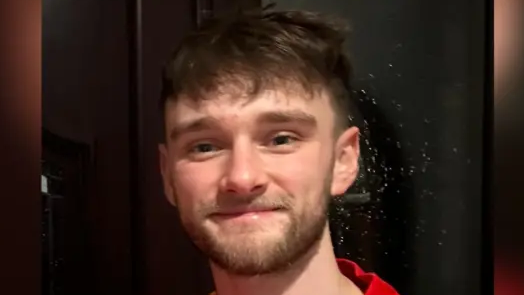 Kieran Hughes is a young man with thick eyebrows and a short beard. He has short brown hair which is shaven at the sides and is wearing a red top. He is smiling