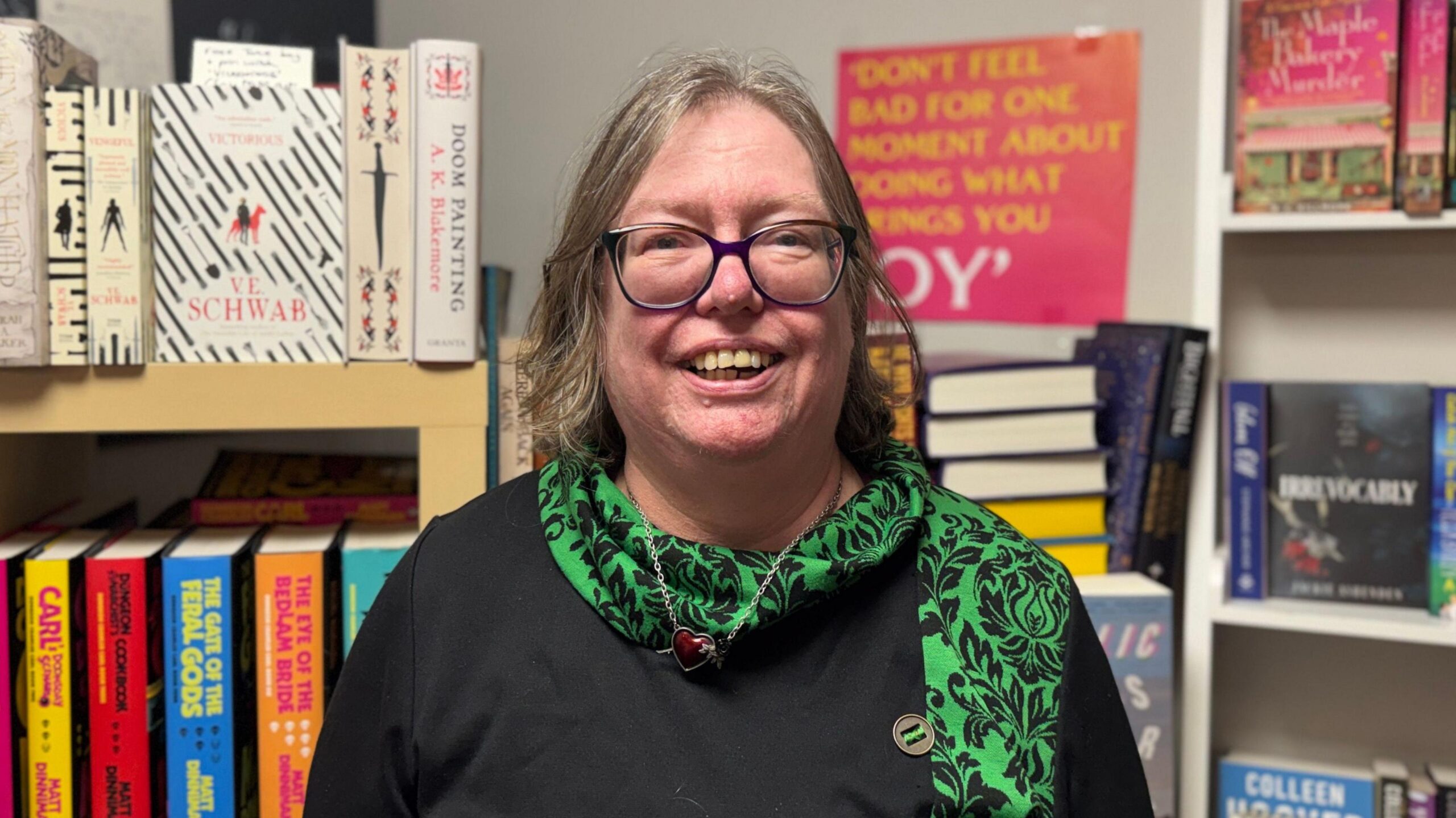 Jo Zebedee smiles at the camera. She has short, greying hair and is wearing a pair of glasses, as well as a green patterned scarf and a black shirt. She is standing in front of a bookshelf.