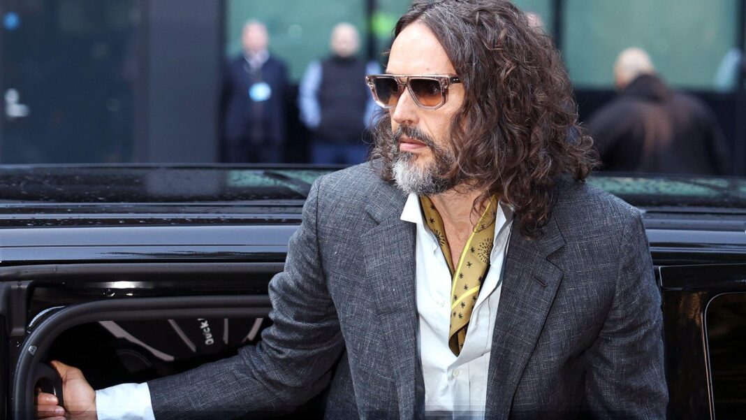 Woman says she ‘closed eyes and tried not to be there’ during alleged Russell Brand rape Russell Brand gets out of a car wearing brown sunglasses, a grey suit with a yellow fabric around as his neck. He has long hair down to his shoulders.