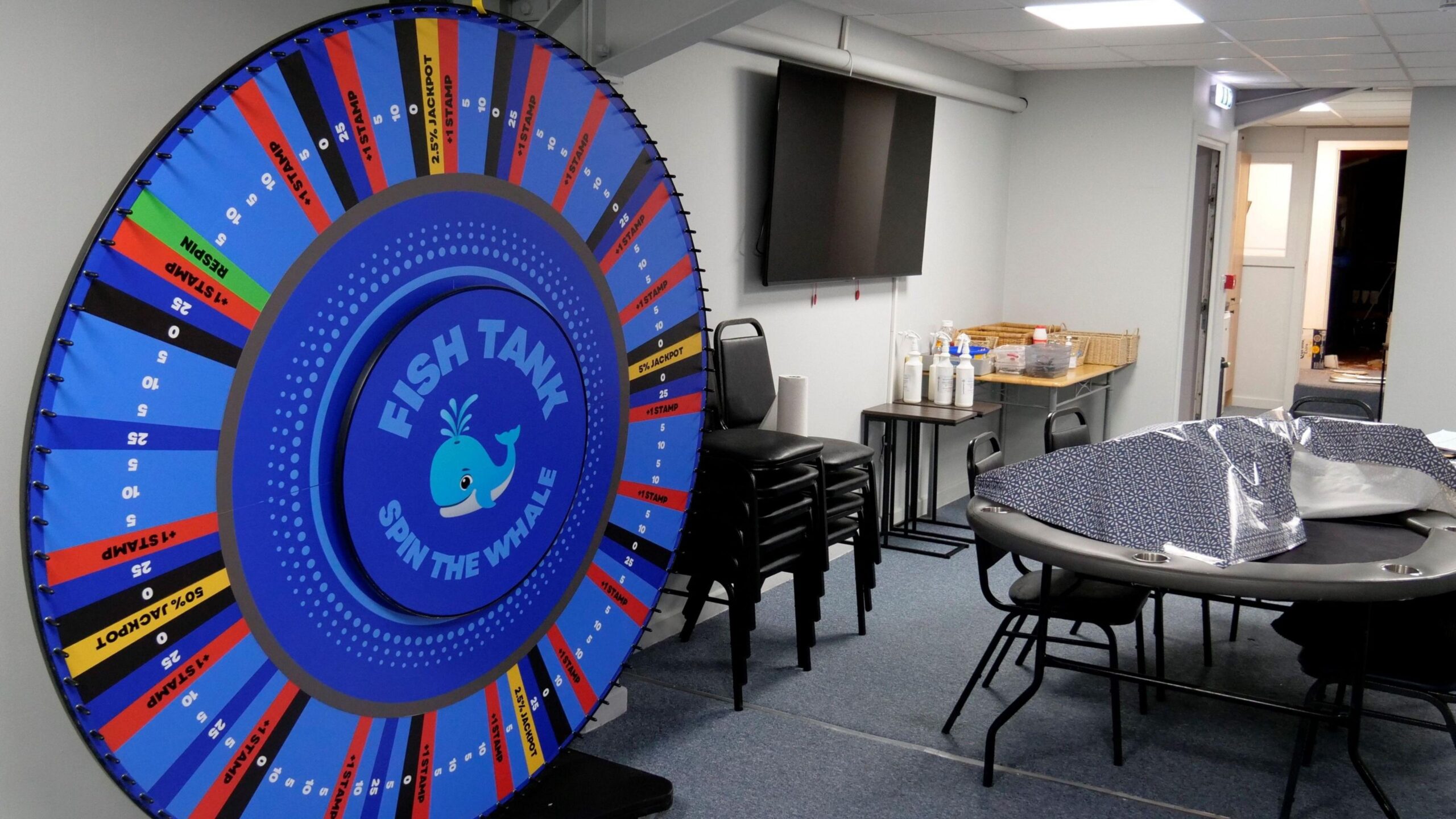 An office-type room with a large, multicoloured prize wheel erected against one wall on the left, with a gambling table surrounded by chairs in the middle of the room. More chairs are stacked up against the wall and there is a TV mounted on the same wall.