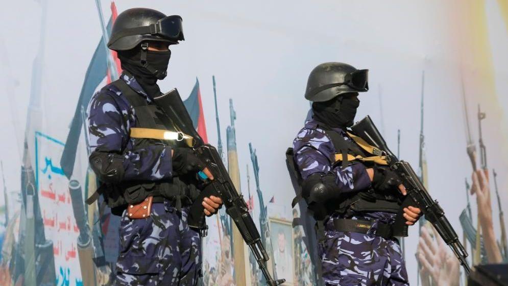 Two soldiers with guns in blue camouflage standing guard in front of a big mural that has may pictures of guns on it.