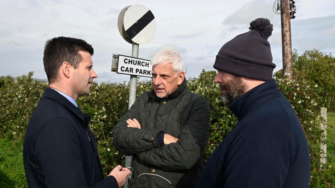(Left to right) DUP MLA Jonathan Buckley, Nigel Dawson, the district master of the Orange Order in Portadown, and deputy district master Alastair Power. Johnathan has short dark hair, wearing a navy jacket. Nigel has short white hair, wearing a khaki rain jacket with his arms folded. Alastair has a dark beard, wearing a black bobble hat and navy fleece. They are all standing in a circle. A sign for the church car park and shrubs are behind them.