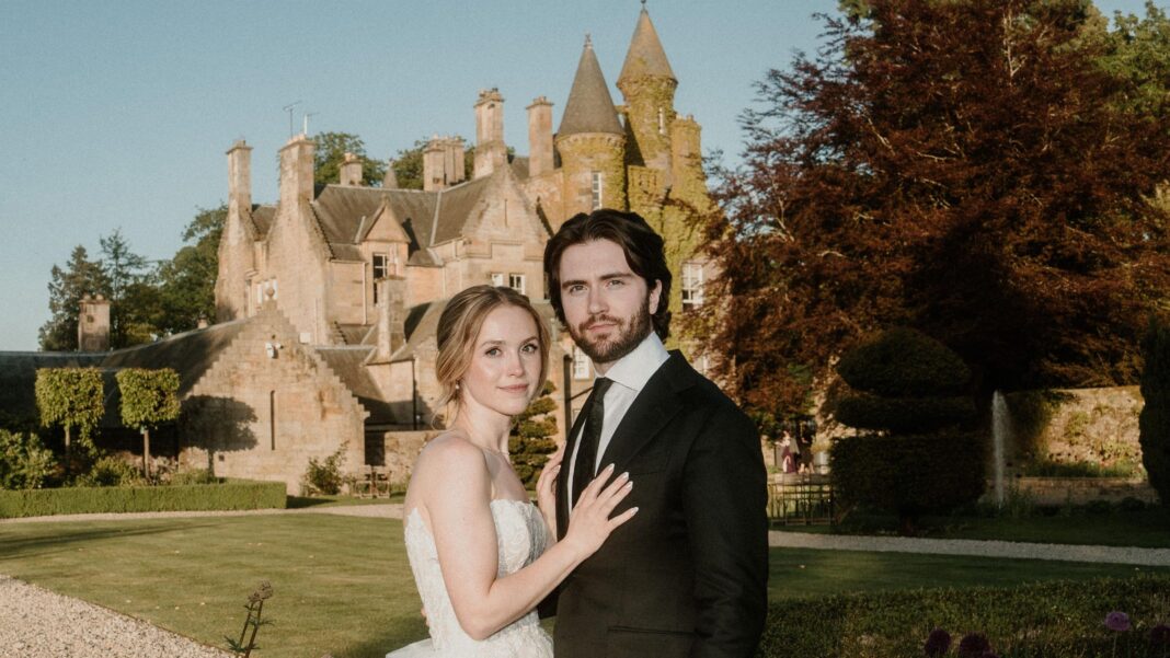 Why thousands of couples are choosing Scotland to say ‘I do’ Savannah who is blonde, fair skinned and wearing a white wedding dress stands next to her husband Hayden, who has dark hair and a beard and wearing a black suit. They stand in front of a Scottish Castle with the sunshining.
