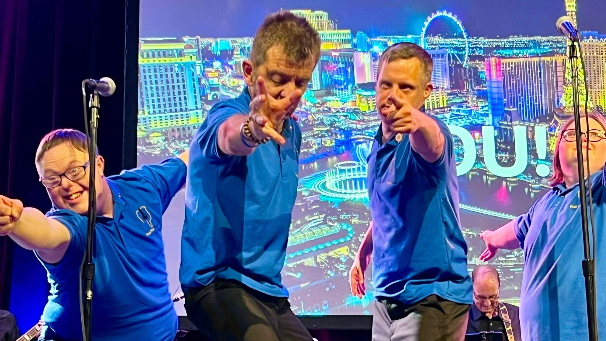 Four performers in blue tops with microphone stands pointing towards the camera which is in the audience.