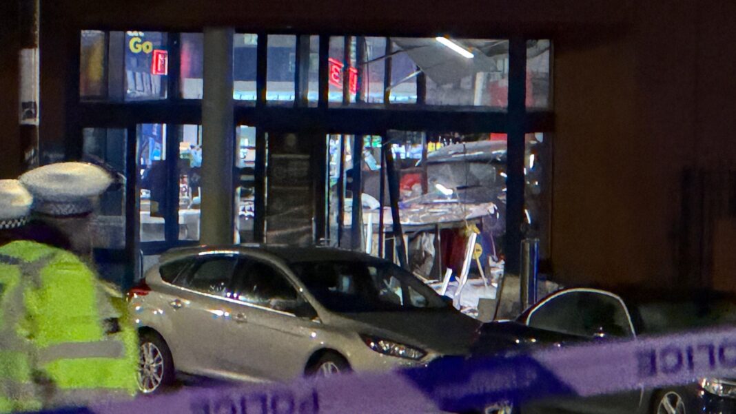 A police officer stands in front of a silver car which is next to a glass door to a supermarket.