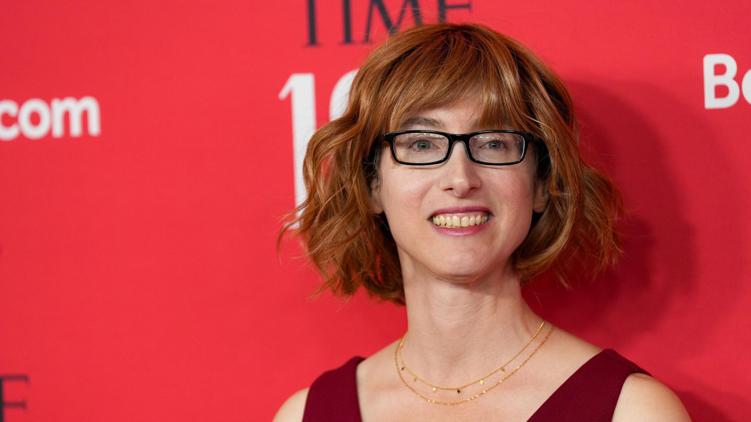 Freida McFadden standing in front of red, black and white background with glasses on