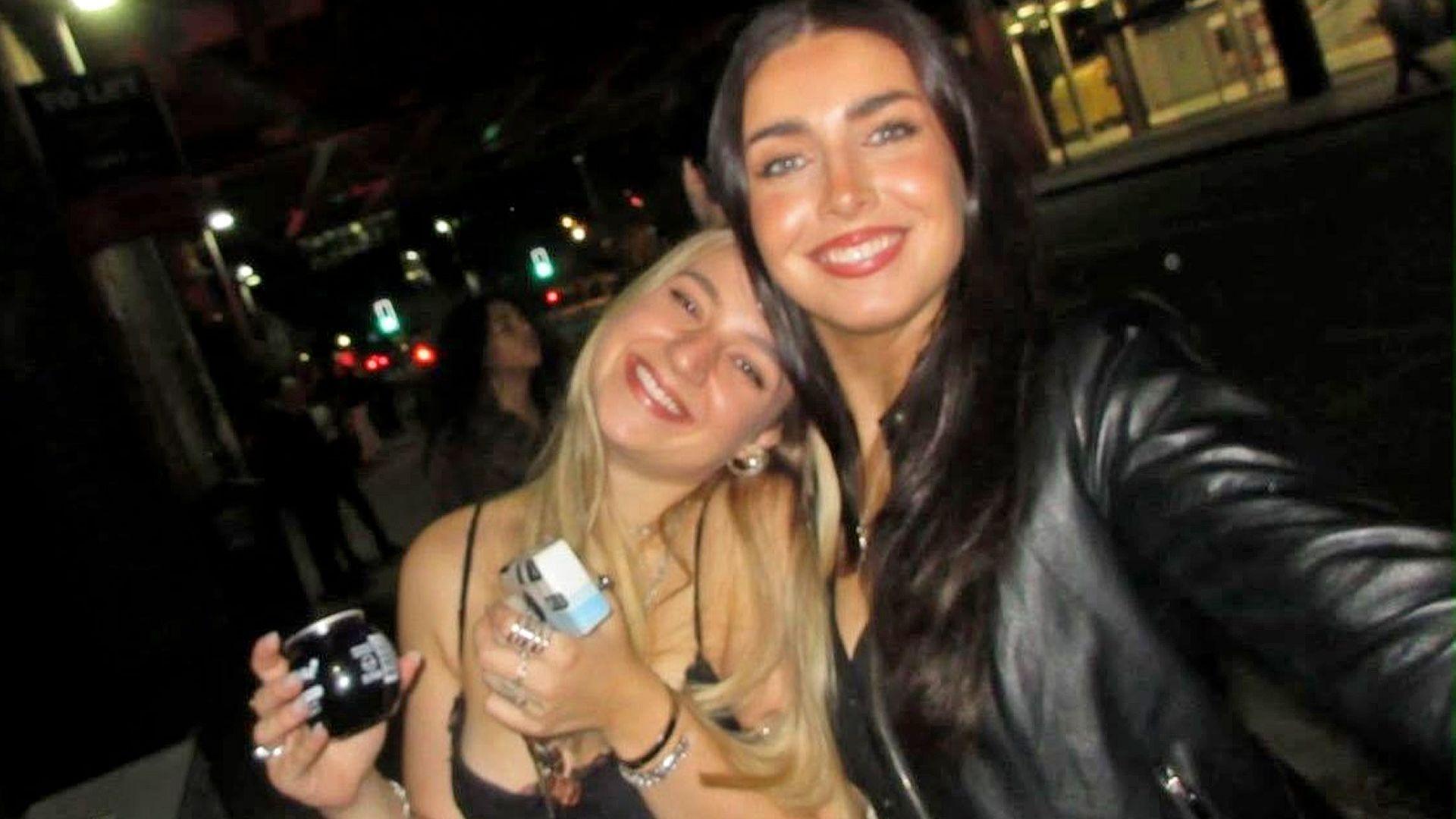 There are two girls in a dark bar smiling at the camera . On the left a blonde girl is wearing a strappy top and holding a drink. She leans into her friend on the left who is holding the camera to take a selfie of the two of them. The girl on the right has long brown hair and is wearing a black leather jacket.