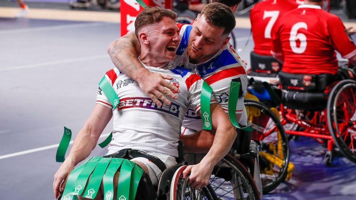 Rob Hawkins, seating in a wheelchair in a white kit with green tags on his shoulders and knees, is hugged from behind by teammate Tristan Norfolk