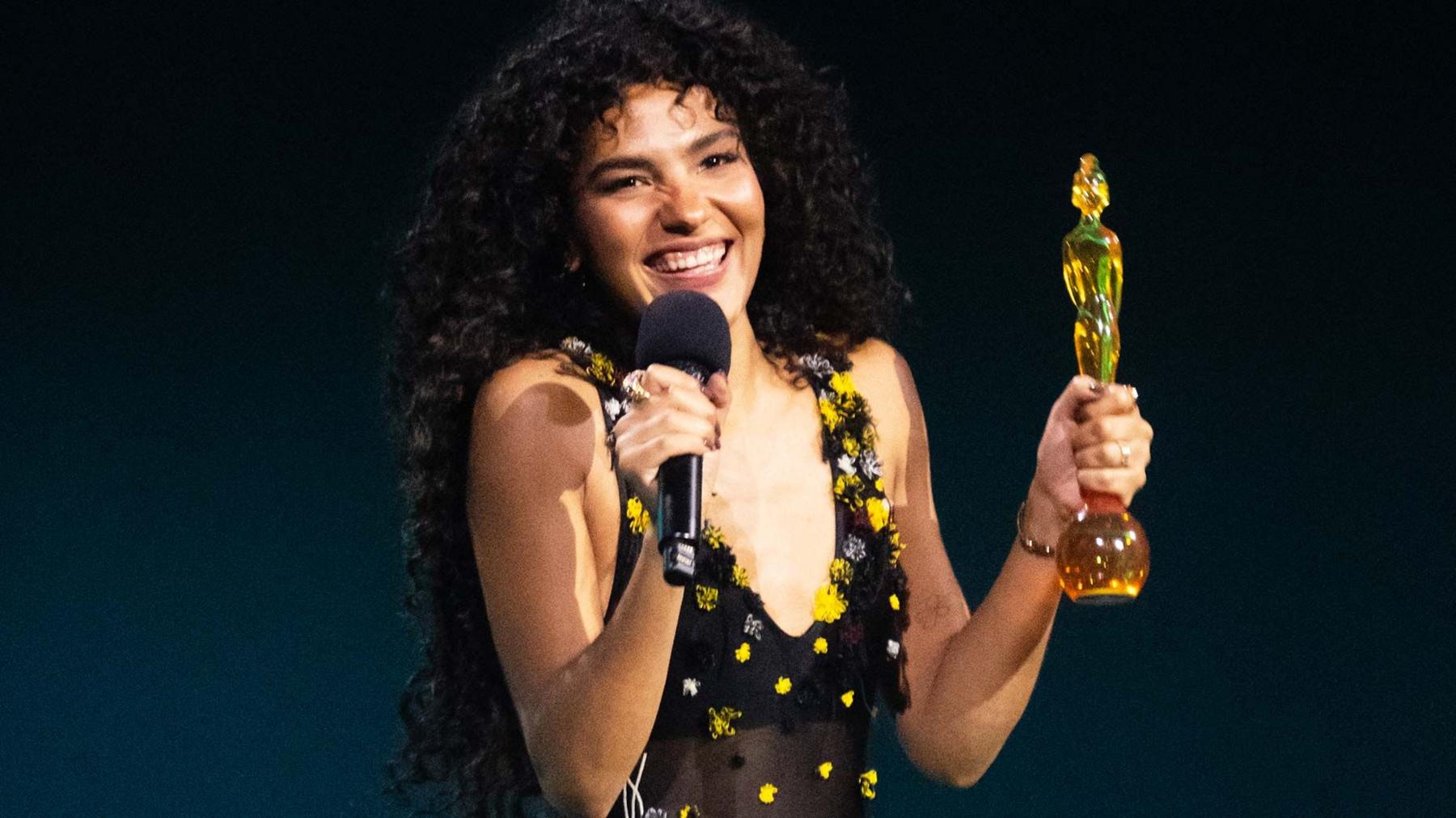 Olivia Dean smiling holding a brit award trophy at the 2026 Brit Awards. She is wearing a black dress with yellow spots