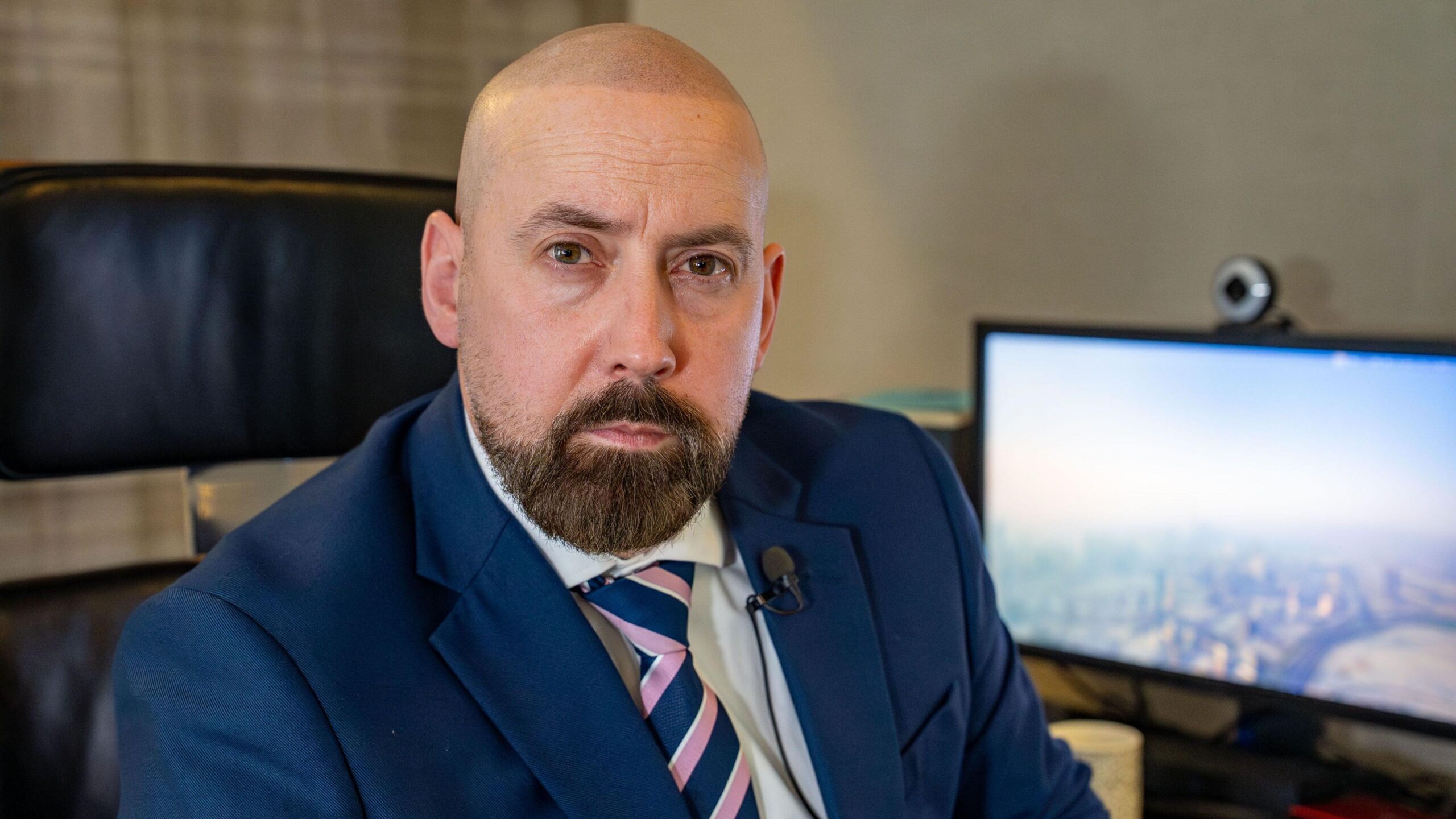 David Kennedy looking at the camera. He is wearing a blue suit jacket over a white shirt with a blue and pink striped tie. He is sitting on a chair in an office and a computer screen and webcam can be seen in the background.