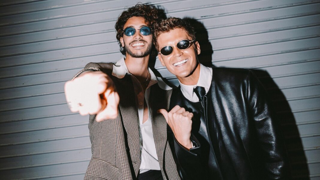 Babydoll singer says life will ‘never be the same again’ after UK number one Two men stood next to each other. One is pointing at the camera wearing a suit jacket, open buttoned shirt and blue tinted glasses. The other wears a leather jacket, shirt. tie and black tinted glasses.