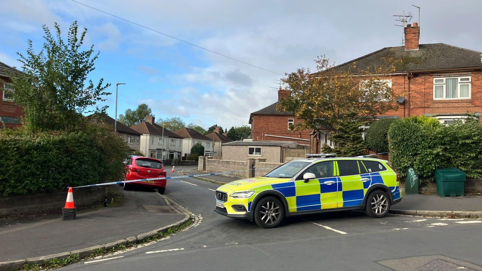 A marked police vehicle blocks the entrance to a residential street that has been sealed off with police tape and traffic cones. Semi-detached houses and parked cars line the road, while the cordon extends further down the street under a cloudy sky.