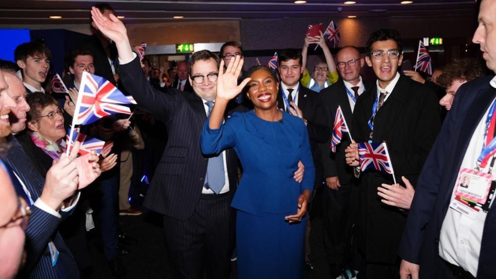 Hamish Badenoch and Kemi Badenoch are surrounded by Tory members, some of whom are holding tiny miniature Union flags. Hamish and Kemi Badenoch are both waving. Hamish is wearing a dark-coloured suit, white shirt and blue tie, while Kemi is wearing a blue dress.