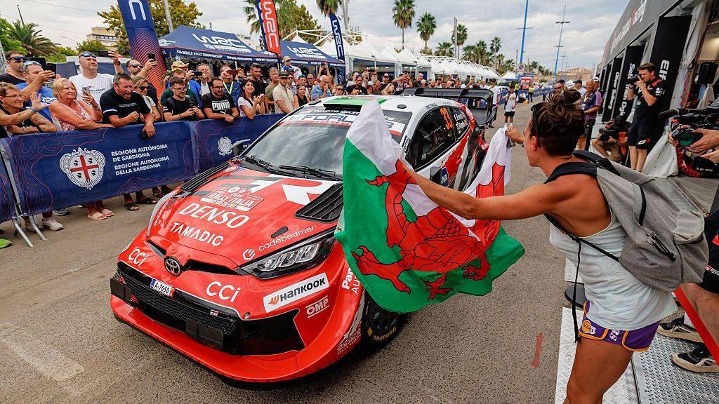 A fan waves a Welsh flag in front of a rally car while fans behind cheer