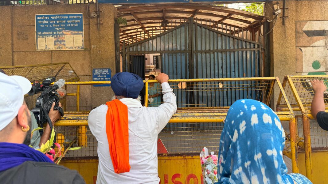 Scottish Sikh activist Jagtar Singh Johal freed on bail after nine years in Indian prison A man wearing a long white garment and a dark blue turban with an orange garment slung over his left shoulder stands against a yellow fence in front of prison gates in Delhi, India. The back of a woman's head, who can be seen clutching flowers, and a man in a white cap are in the foreground.