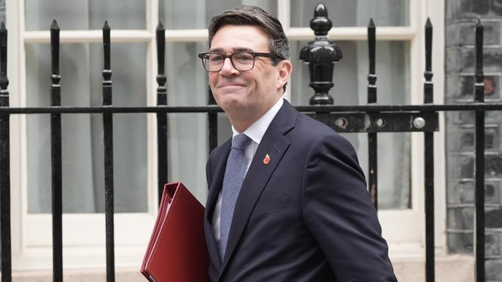 Andy Burnham smiles outside the railings of Downing Street while holding a red folder and wearing a suit.
