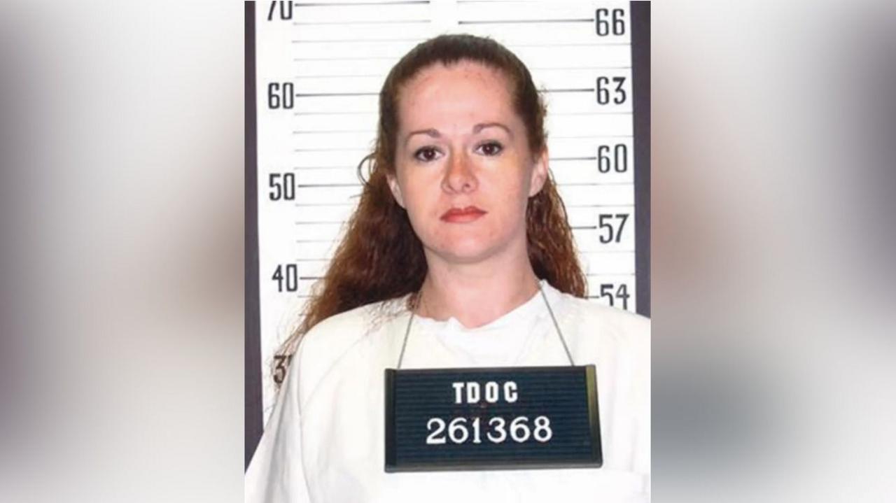 A woman with red hair stands in front of a sign that shows a person's hight, she wears a sign around her neck reading "TDOC 261368"