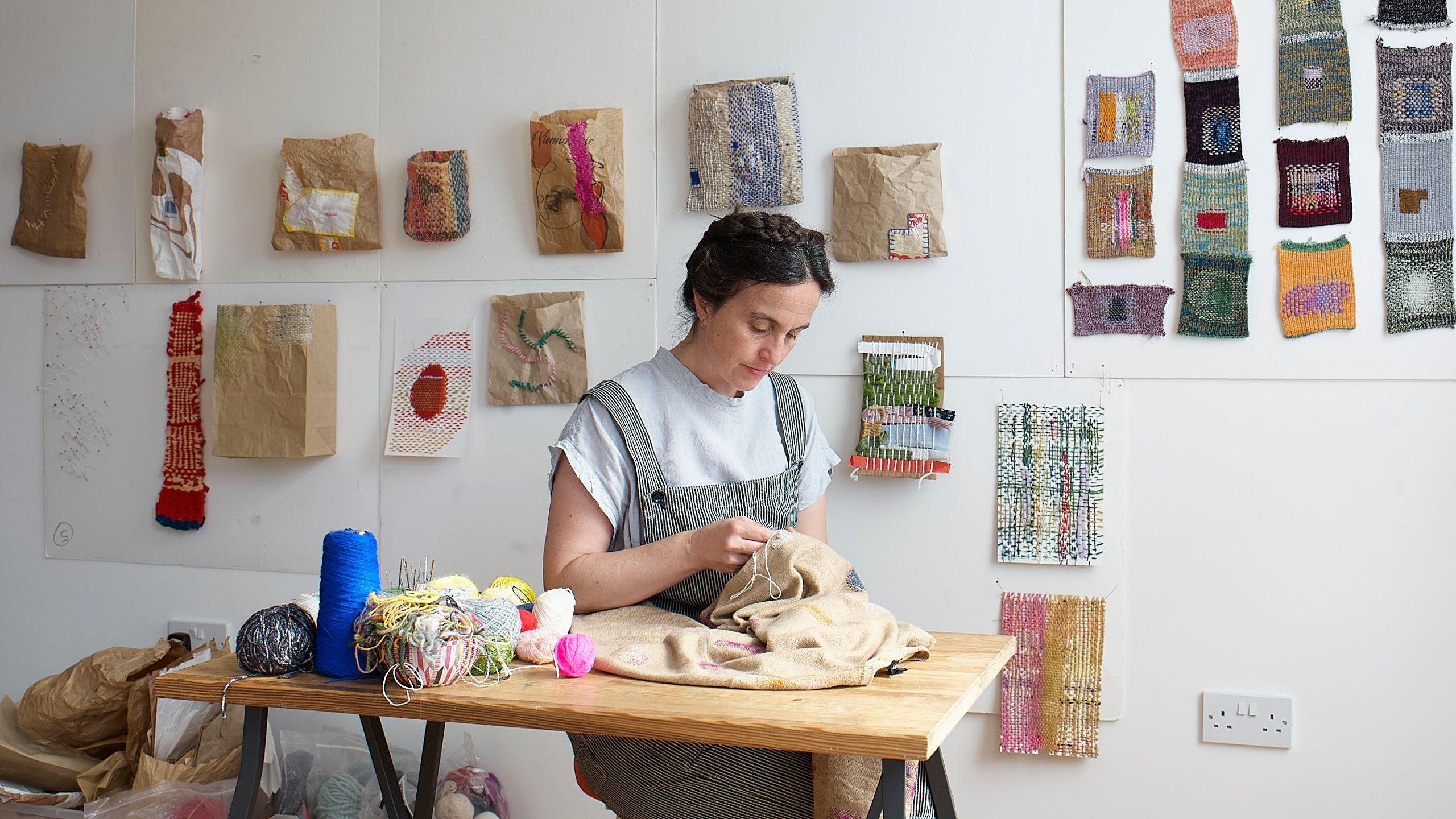 Woman mending at a table with samples on the wall behind her