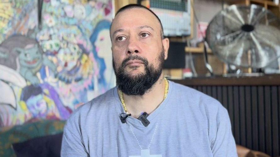 Dr Hakeem Stevens, who is wearing a pale blue long-sleeved T-shirt, sits in a room. There is colourful art on one of the walls behind him.
