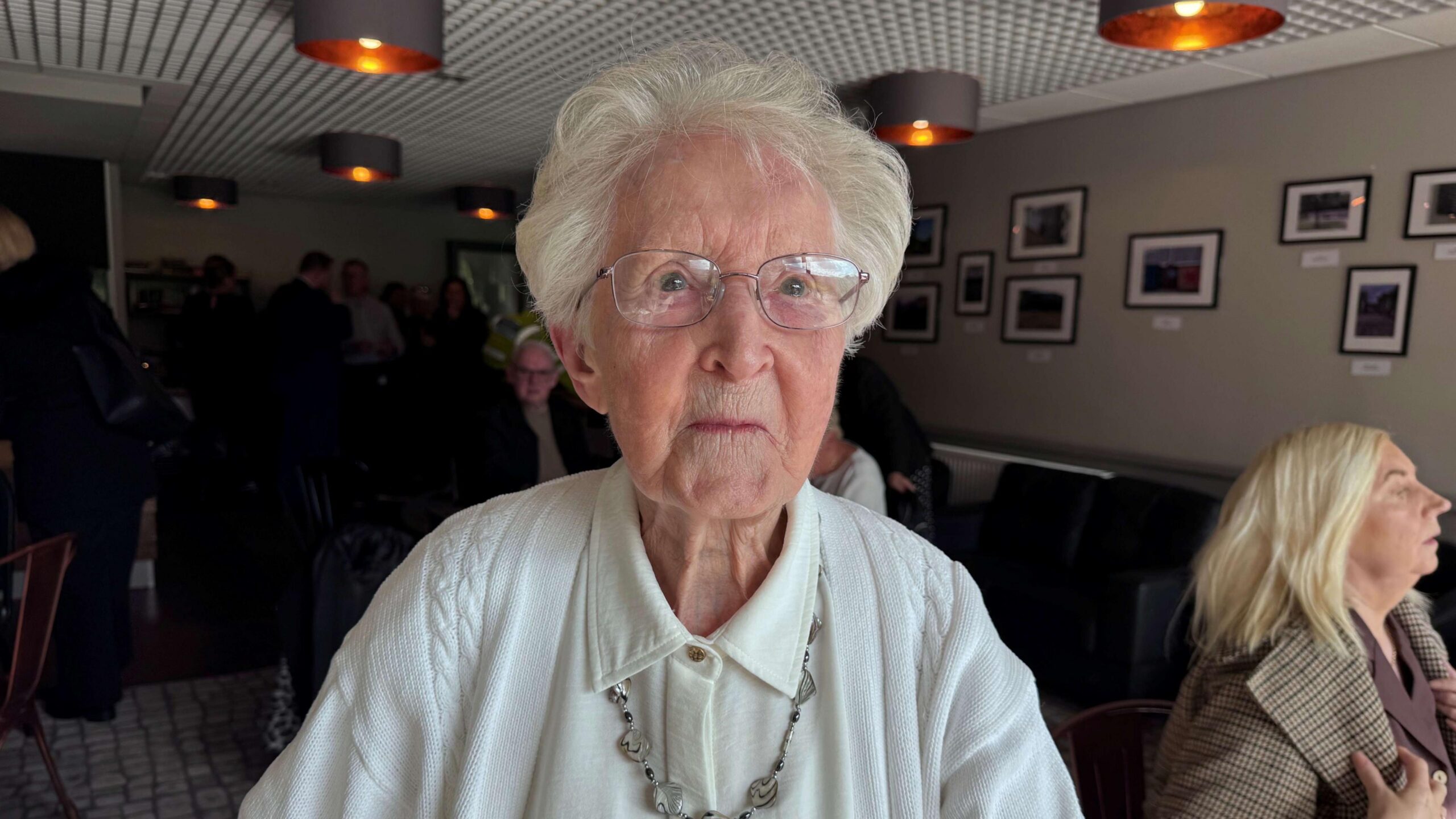 A woman with white hair and glasses looking at the camera. She's wearing a white top and cardigan with a long silver necklace.