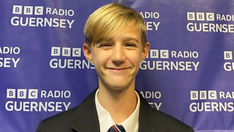 Cole. He is wearing a dark blazer, white shirt and striped tie. He has medium length blonde hair.